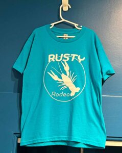 Aqua blue t-shirt with white Rusty Rodeo logo hanging on blue wall 